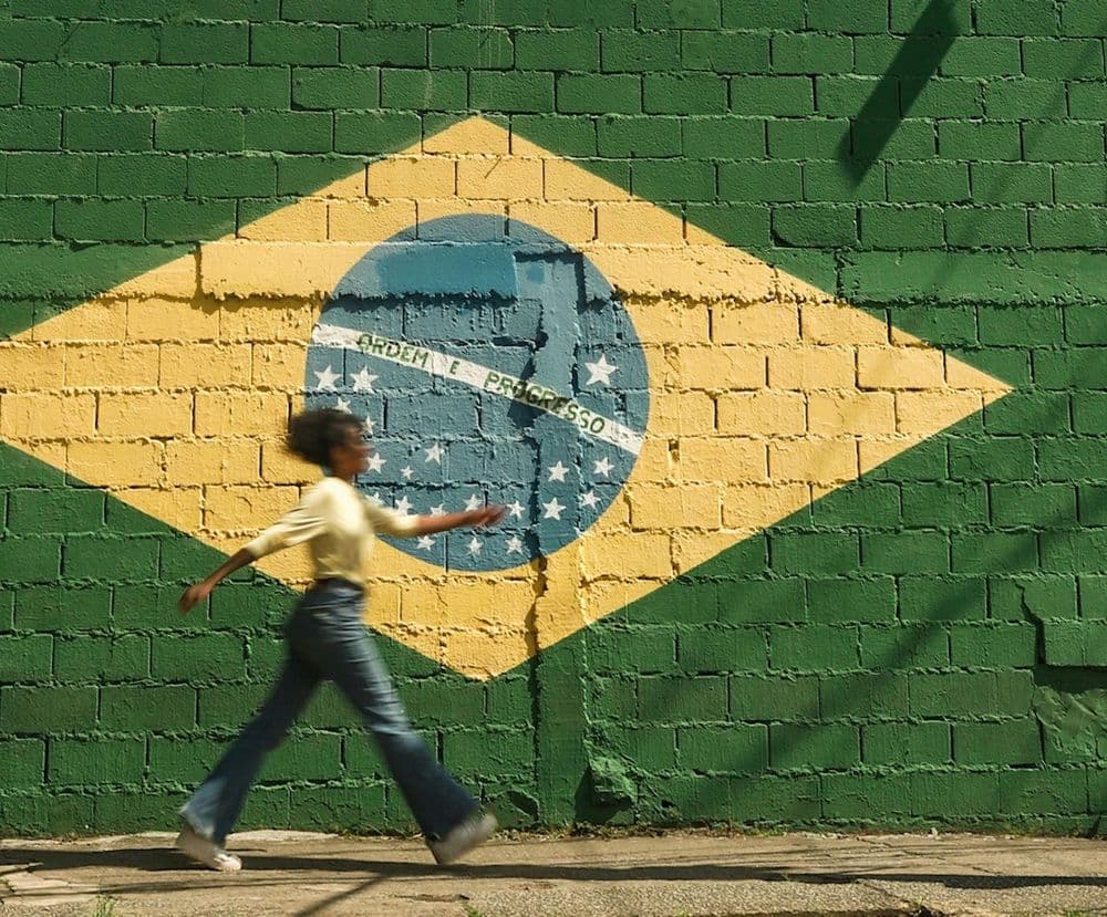 Mulher caminhando em frente a um muro pintado com a bandeira do Brasil
