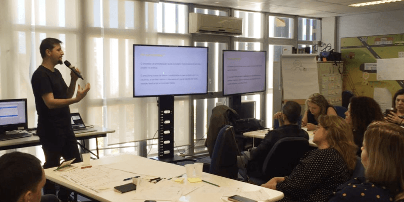 Consultor da Uzzer, Marcon Zanin, de pé em frente a uma tela segurando um microfone e falando para uma turma que está sentada no curso de Cultura UX para o Sebrae Nacional.